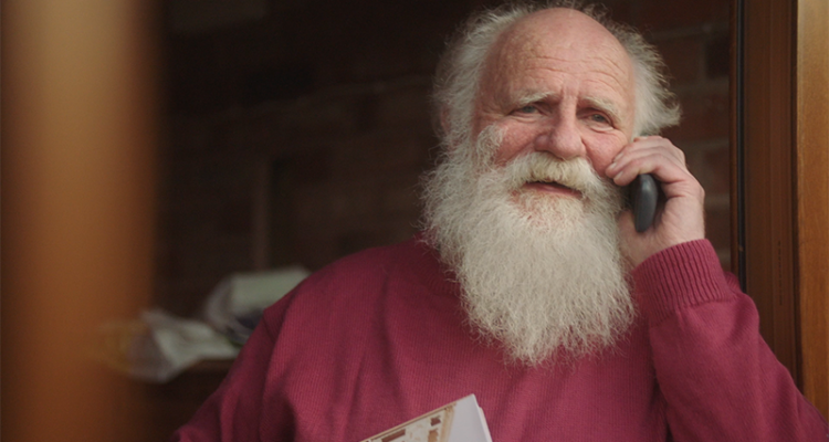 A man with long white hair and a beard holding a phone up to his face on his doorstep