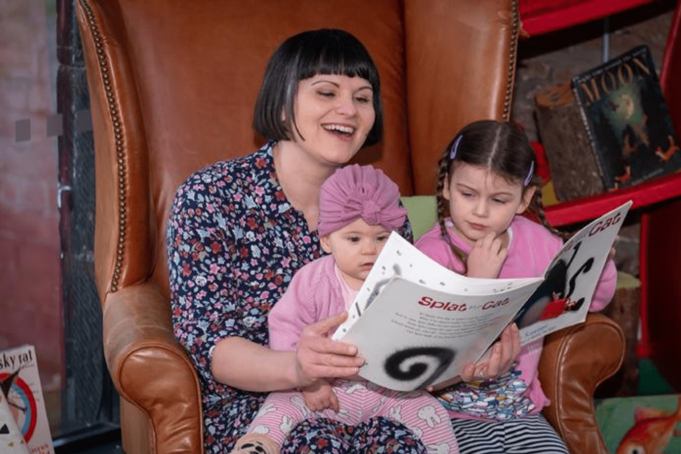 two children sat on the lap of their carer reading a book and smiling