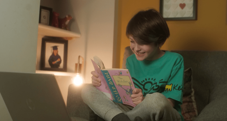Child reads book in front of laptop where his volunteer reads with him