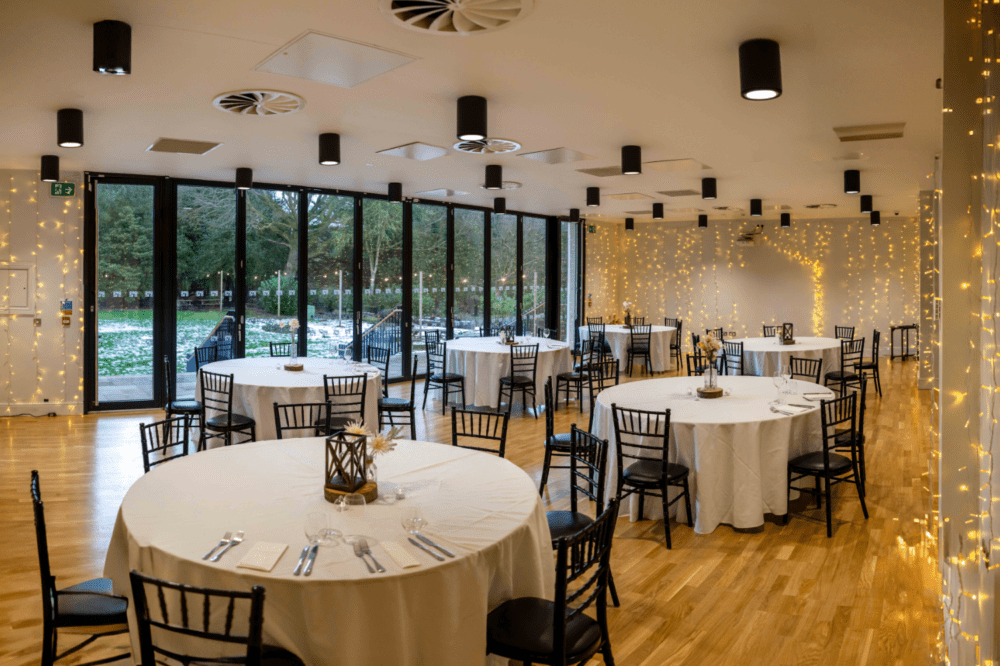 The theatre room dressed with round tables, white table cloths and black chairs. Fairy lights are strung across the walls twinkling and there are bi-fold door onto the garden.